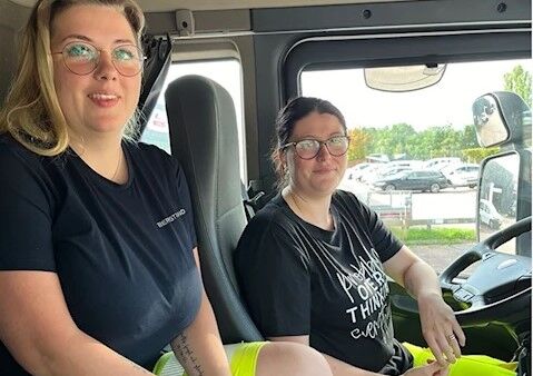 Amanda_Hyckenberg_plus_elev1 Två personer sitter i en lastbilshytt. Den ena sitter på förarsätet och den andra på passagerarsätet. De har båda glasögon och vardagskläder. En parkeringsplats är synlig utanför fönstret.