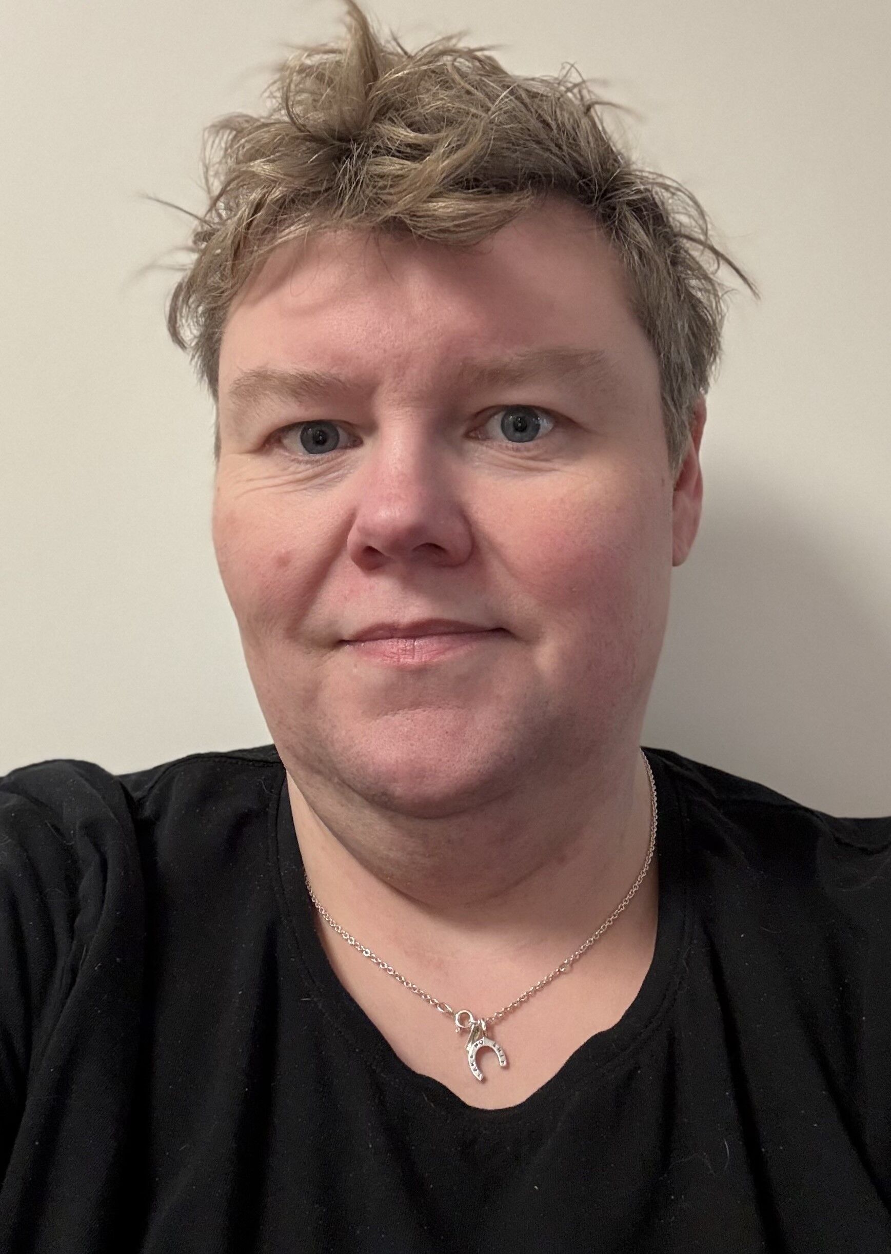 Person med kort hår klädd i en svart skjorta och ett halsband och tittar på kameran mot en vanlig bakgrund.