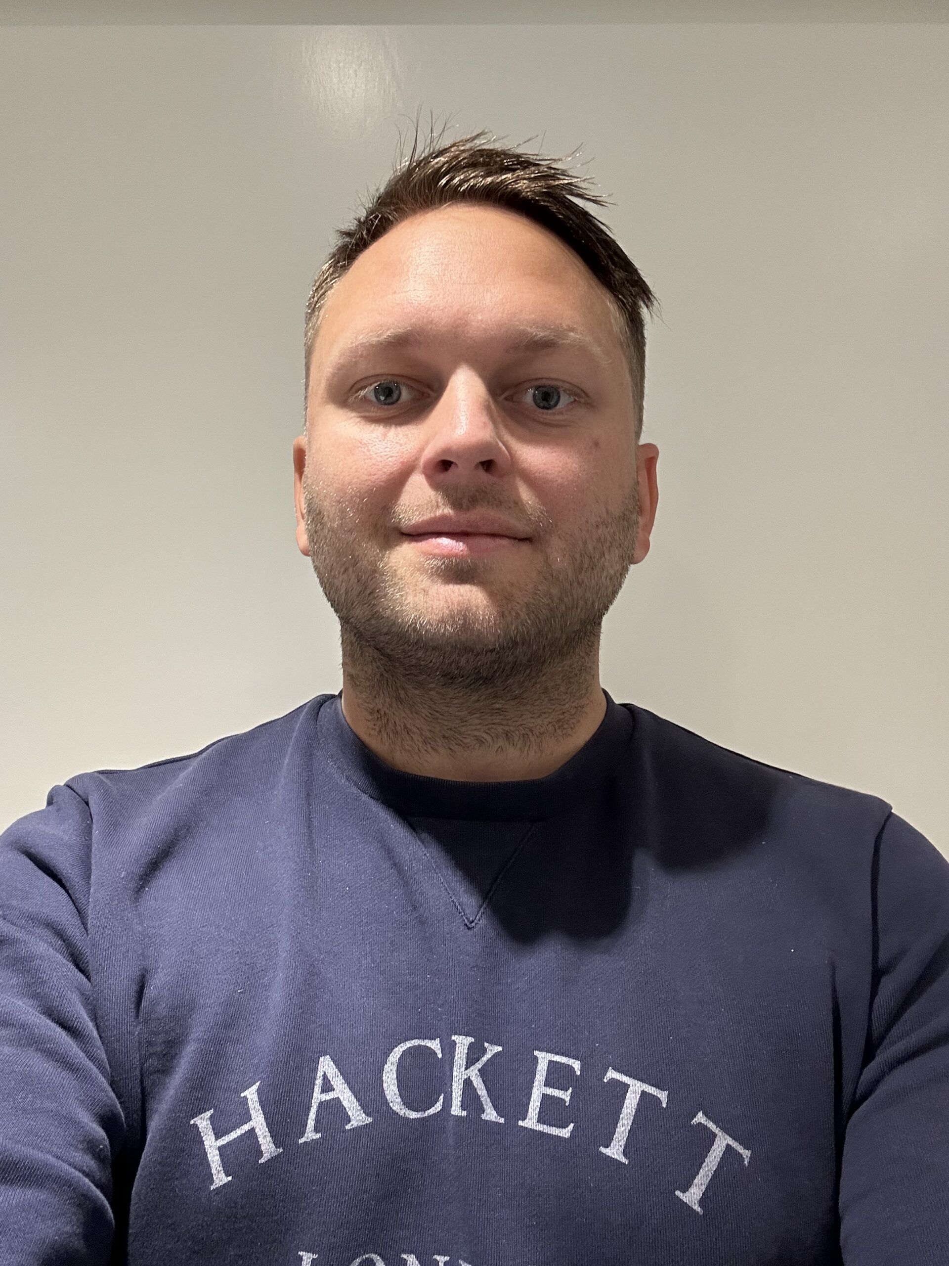 En man med kort brunt hår och en marinblå Hackett London-sweatshirt står framför en vit bakgrund och tittar in i kameran.