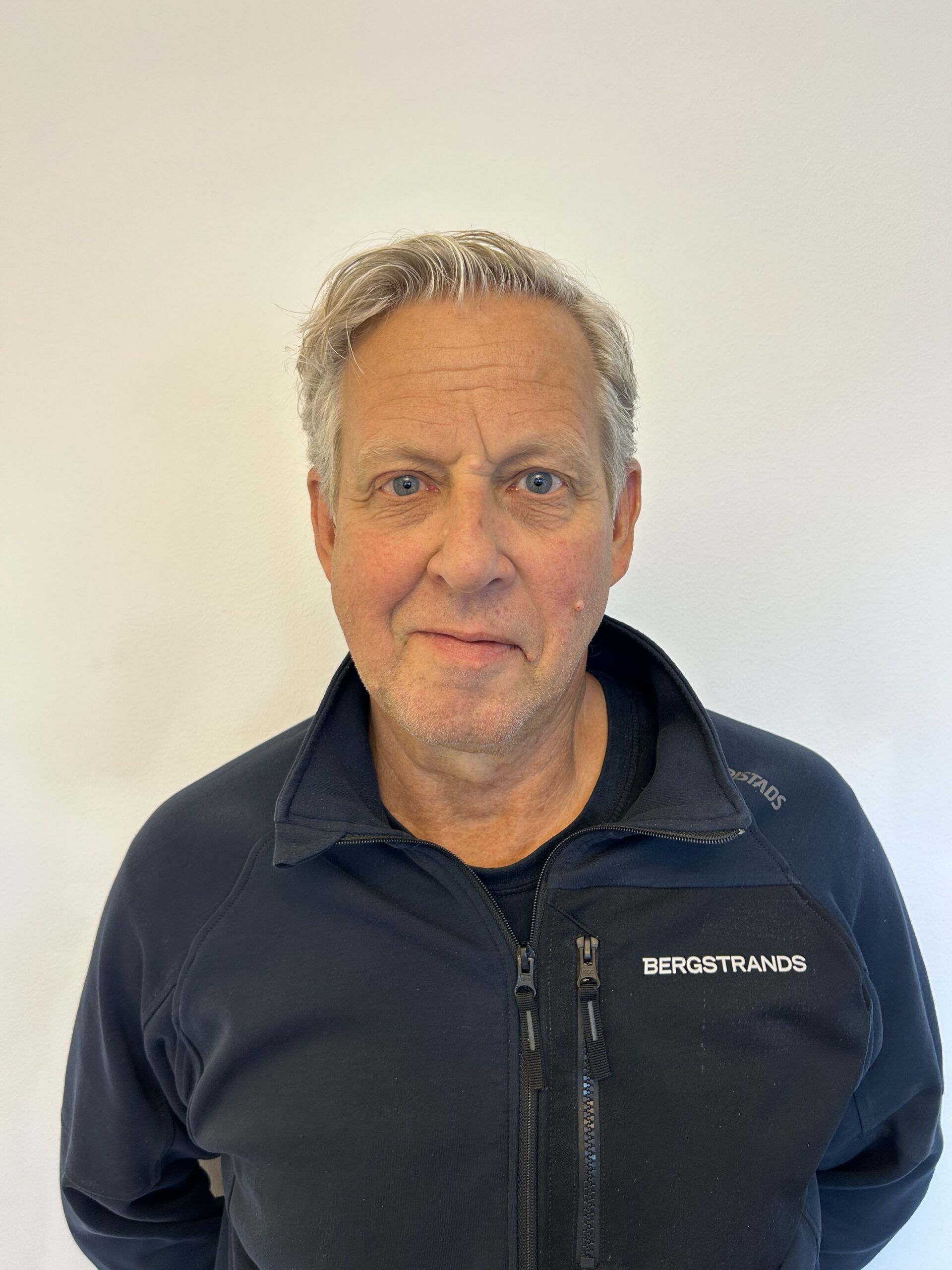 En äldre man med grått hår, klädd i en mörk jacka med dragkedja och ordet "BERGSTRANDS" på, står mot en ljus bakgrund.