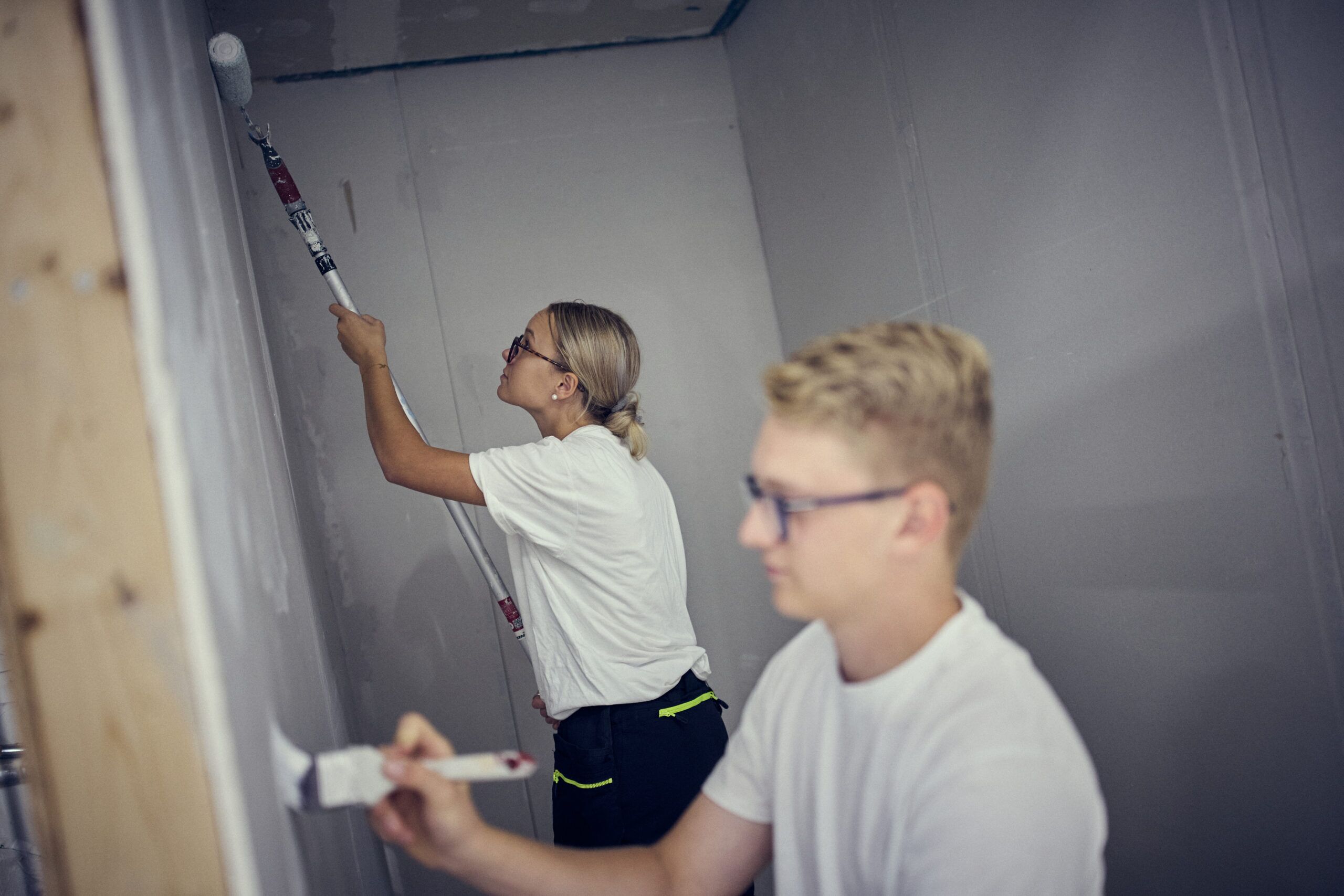 _H8A7987 Två personer i vita skjortor och glasögon målar väggarna i ett rum - den ena använder en roller på taket medan den andra använder en pensel på väggen.